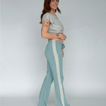 Woman wearing a light gray top and teal pants with white stripes on a plain background