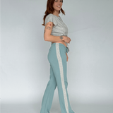 Woman wearing a light gray top and teal pants with white stripes on a plain background